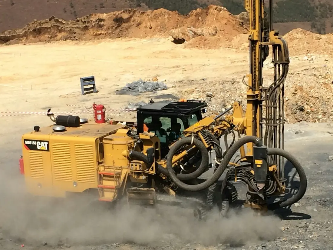 A track-mounted drilling rig working in a cloud of dust on rocky ground