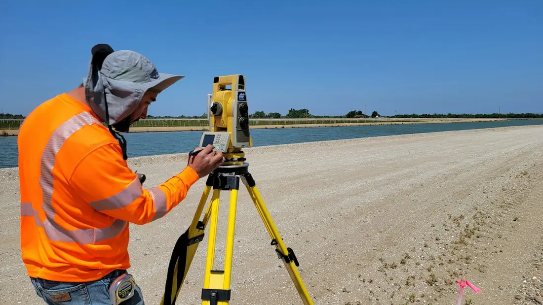 A surveyor in a hi-vis shirt setting up a total station on a tripod beside a canal